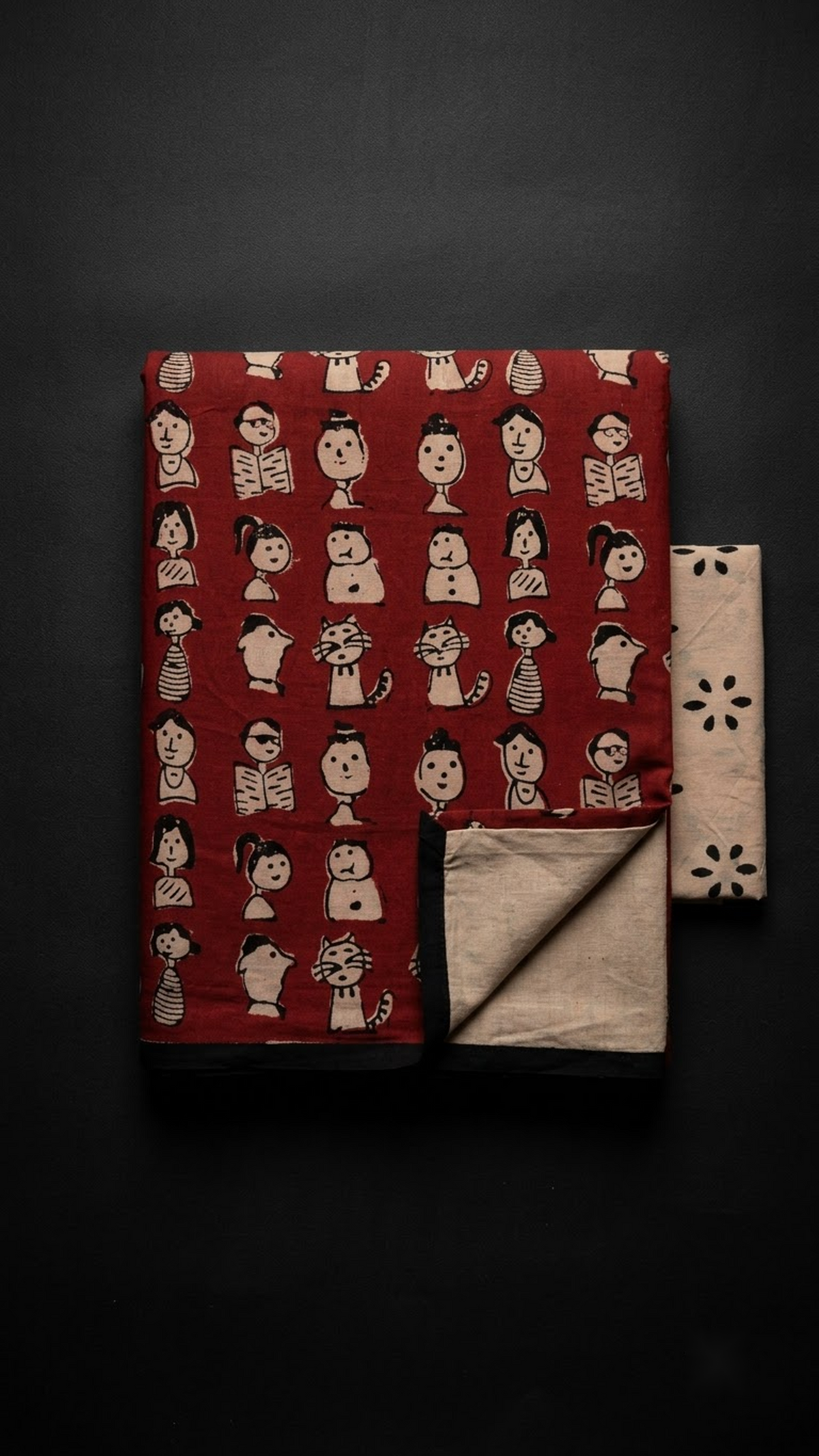Red character print cotton saree with black border and ivory floral blouse, folded on charcoal background