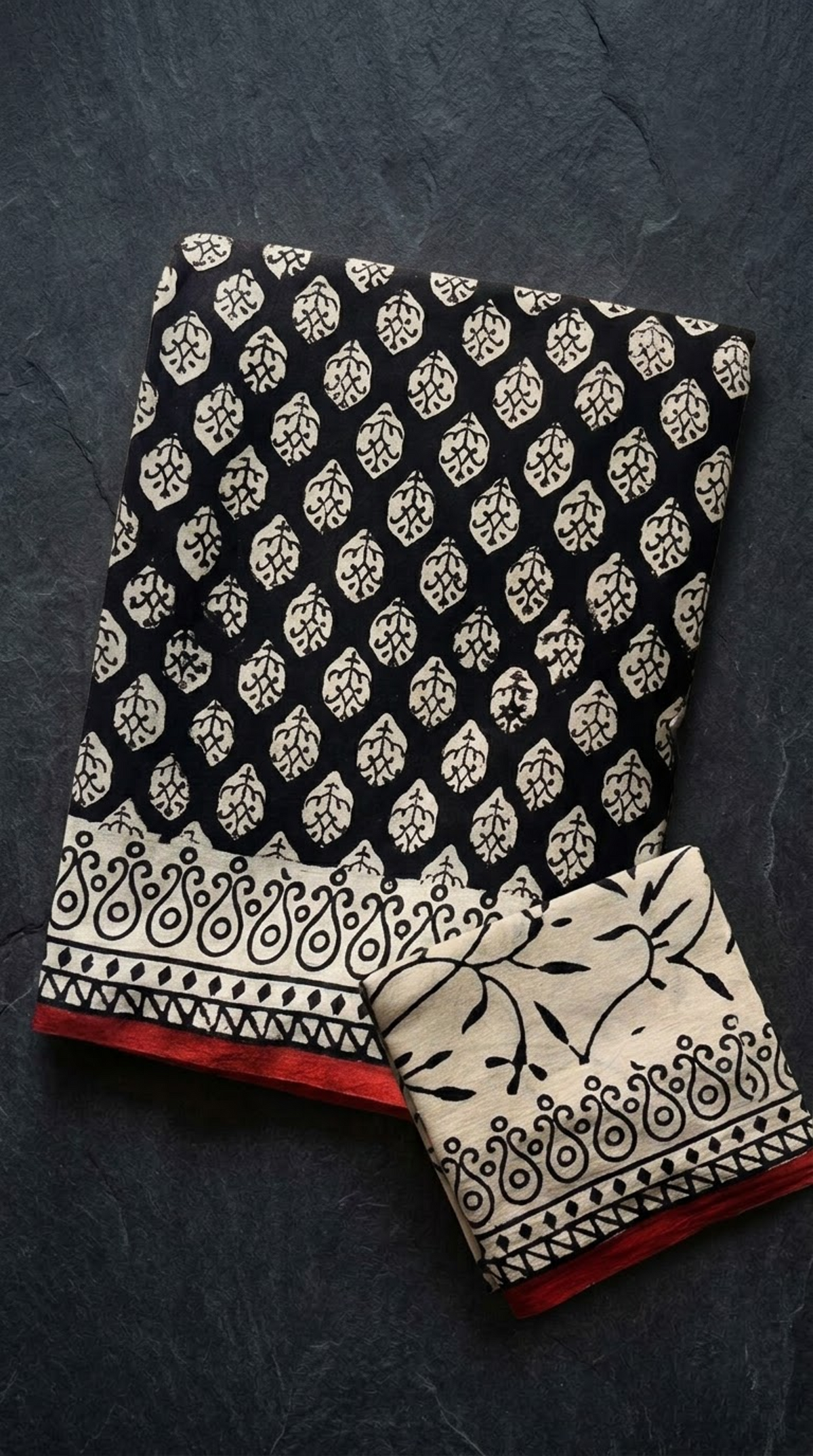 Black cotton saree with ivory leaf motifs, a patterned border with red edging, and an ivory botanical blouse piece, folded on a charcoal background