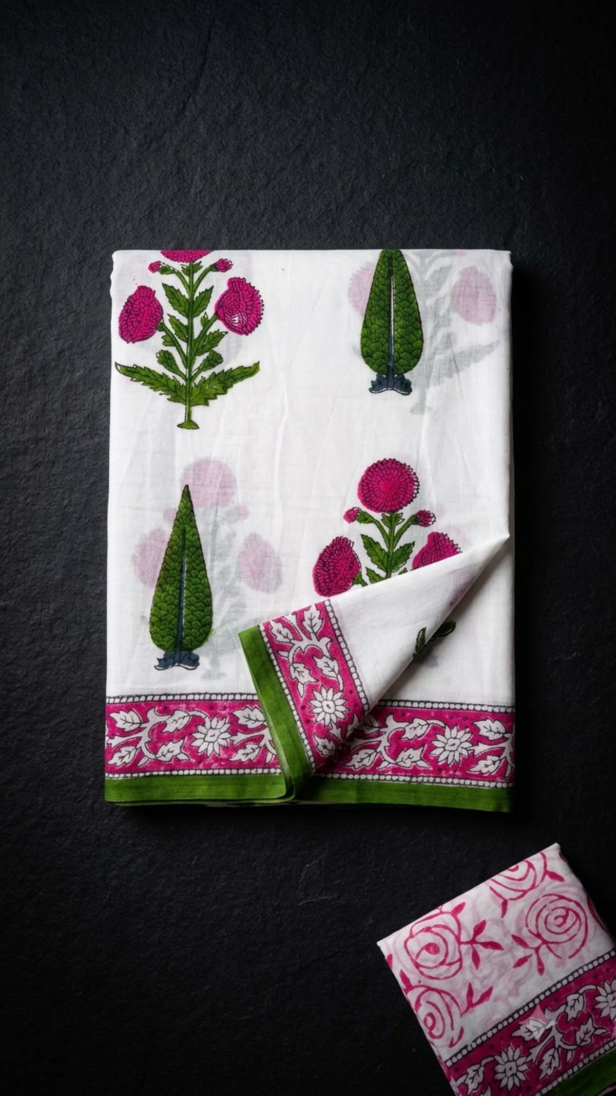 Ivory cotton saree with pink blossoms, green leafy sprigs, pink border, and ivory pink floral blouse piece, folded on a charcoal background
