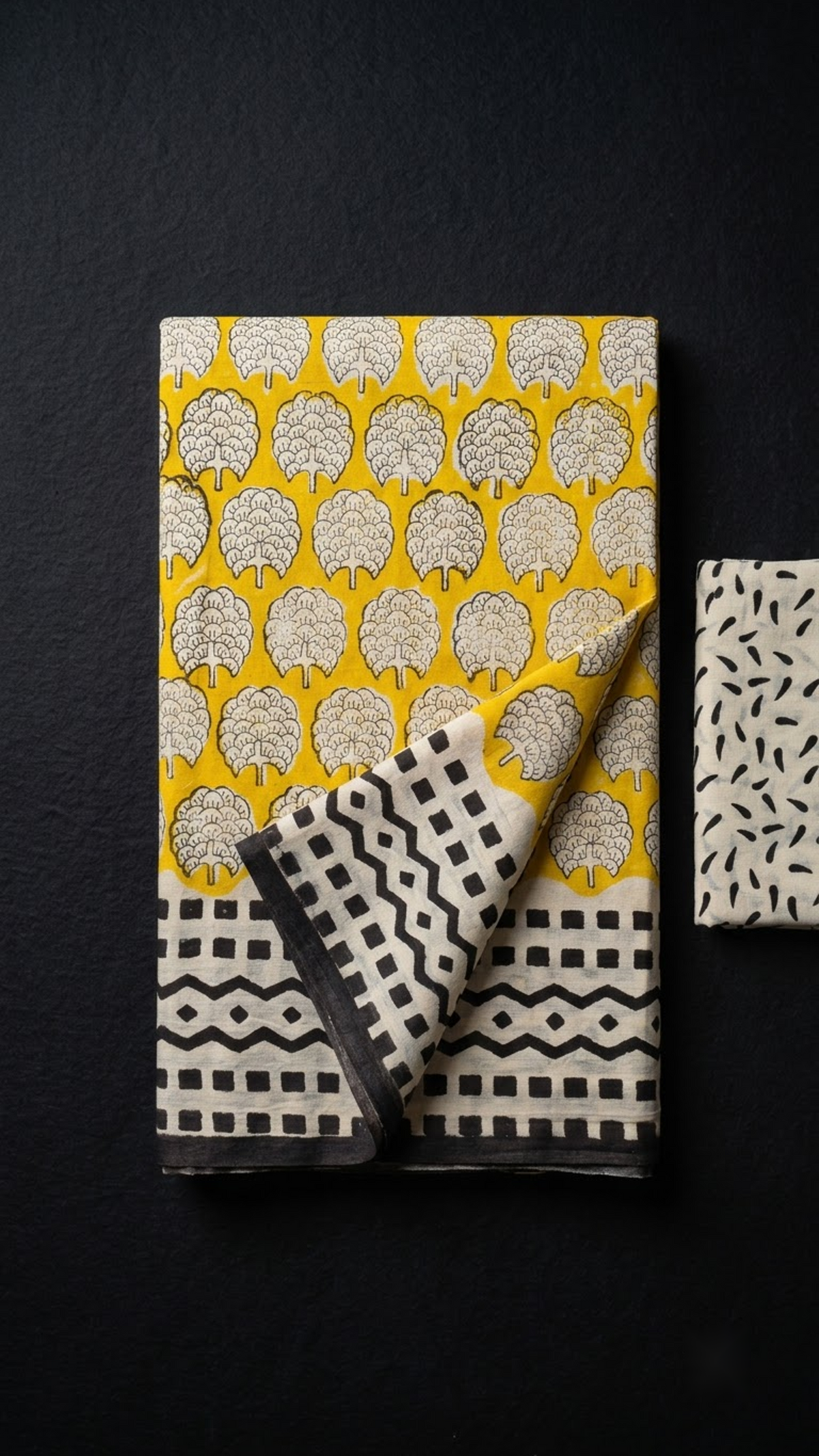 Yellow cotton saree with ivory floral medallions, black geometric border, and ivory black leaf-print blouse piece, folded on a charcoal background