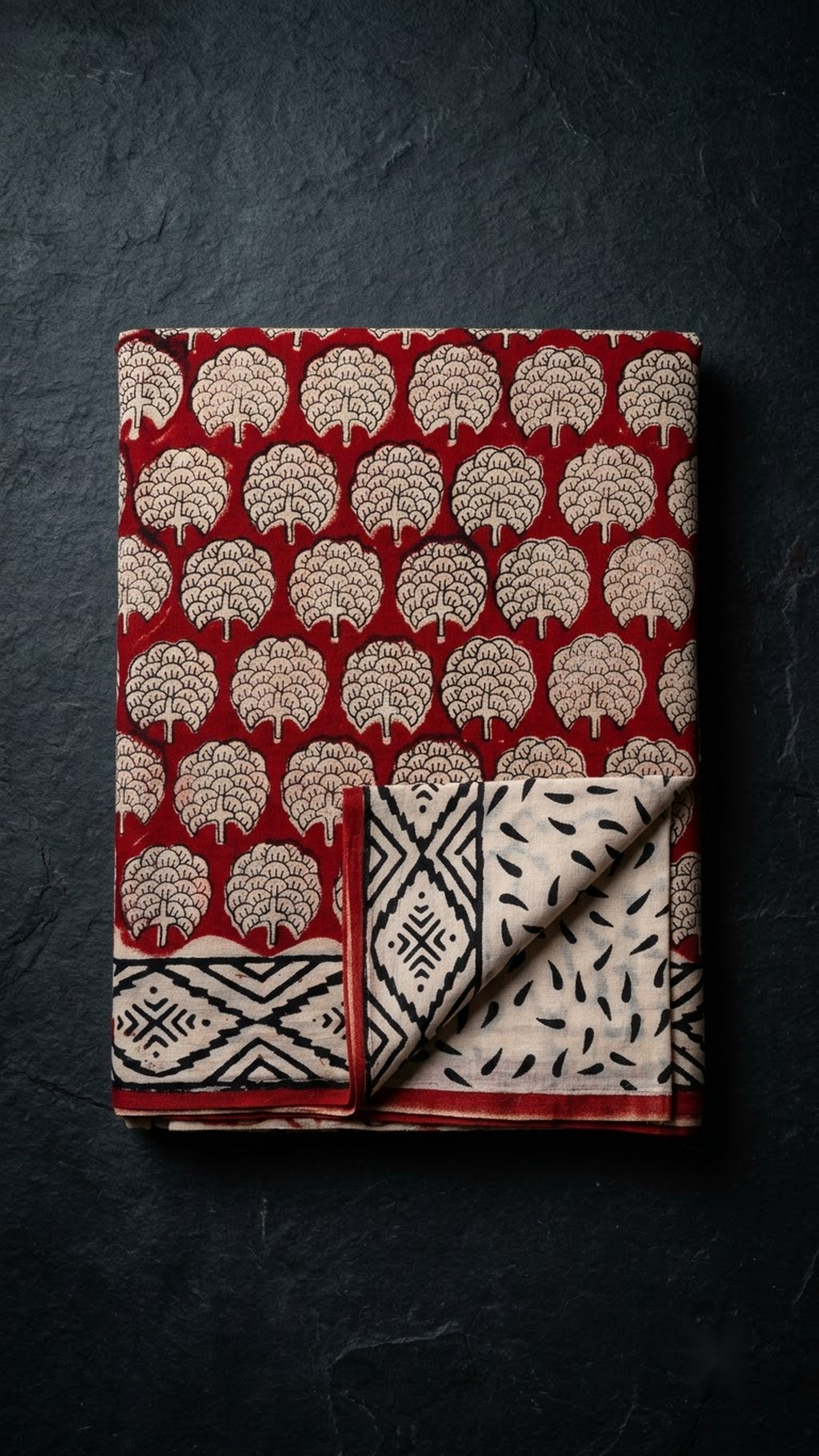 Red cotton saree with ivory floral medallions, black diamond border, and ivory black leaf-print blouse piece, folded on a charcoal background