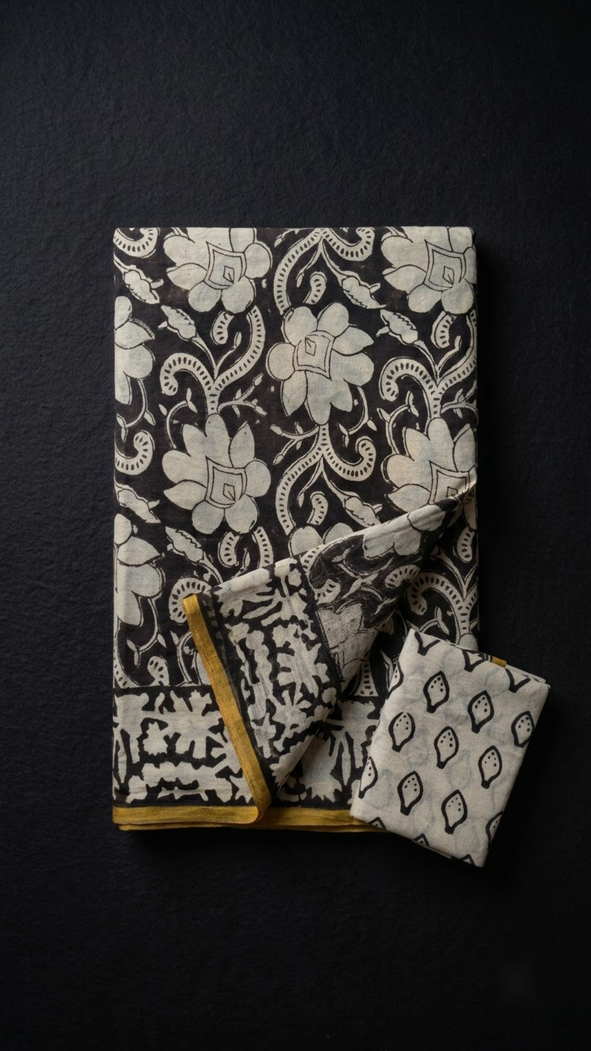 Black cotton saree with ivory floral scrolls, an ivory border with yellow edging, and ivory black motif blouse piece, folded on a charcoal background