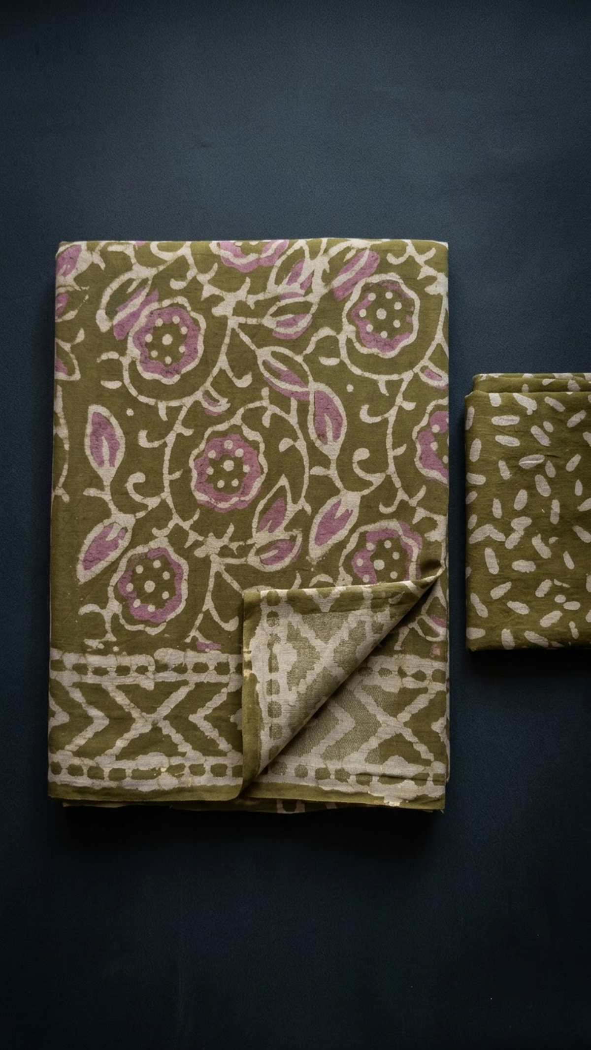 Olive cotton saree with pink flowers and ivory leaves, ivory diamond border, and olive leaf-print blouse piece, folded on a charcoal background