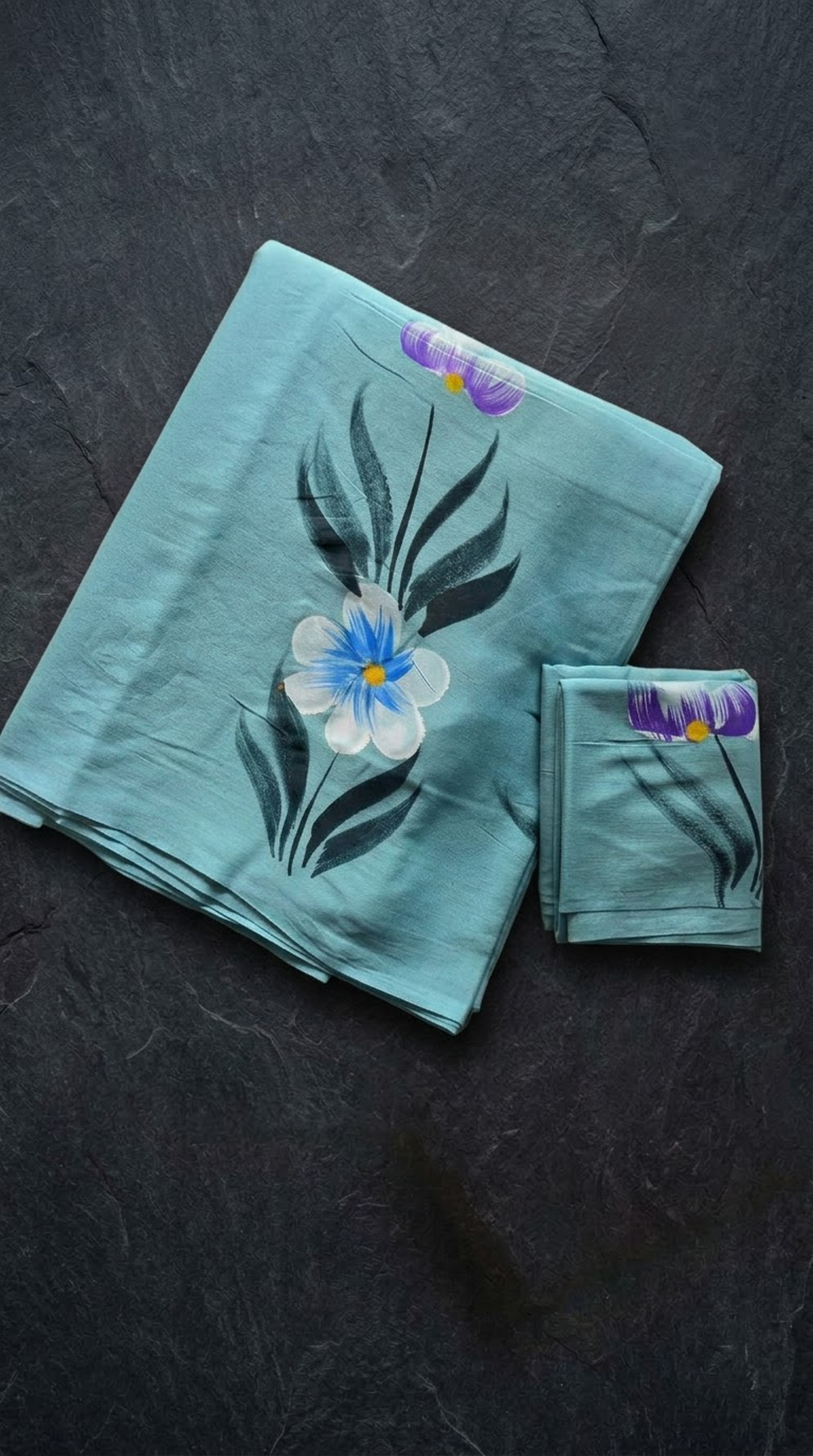Aqua cotton saree with a blue-and-ivory flower, dark teal leaves, purple buds, and coordinating botanical blouse piece, folded on a charcoal background