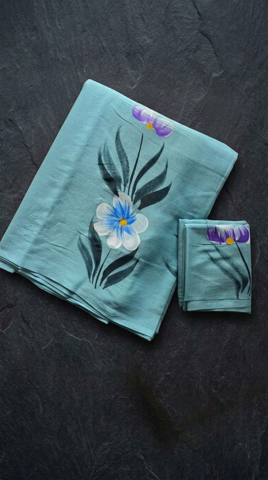 Aqua cotton saree with a blue-and-ivory flower, dark teal leaves, purple buds, and coordinating botanical blouse piece, folded on a charcoal background