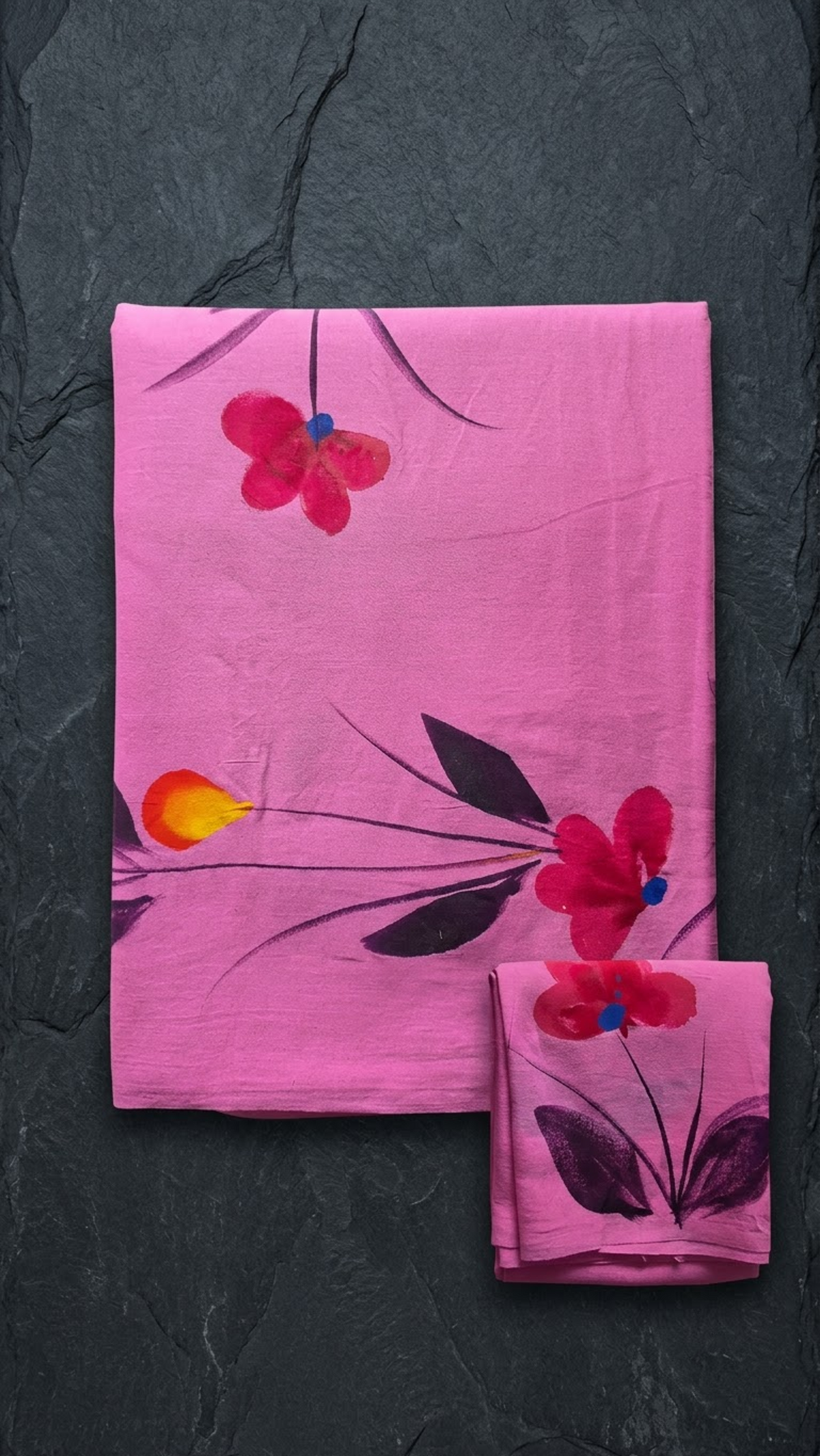Pink cotton saree with red flowers, blue centres, dark leaves, and a matching floral blouse piece, folded on a charcoal background