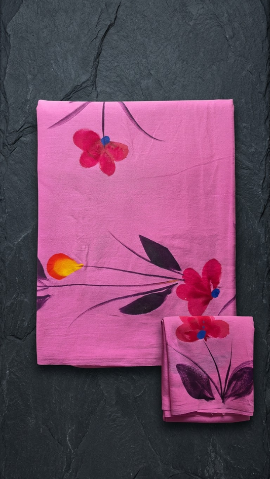 Pink cotton saree with red flowers, blue centres, dark leaves, and a matching floral blouse piece, folded on a charcoal background