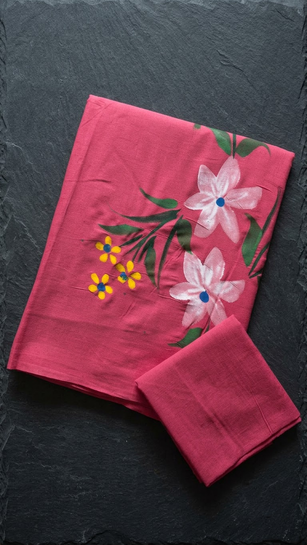 Rose pink cotton saree with white and yellow flowers, green leafy stems, and a plain rose pink blouse piece, folded on a charcoal background