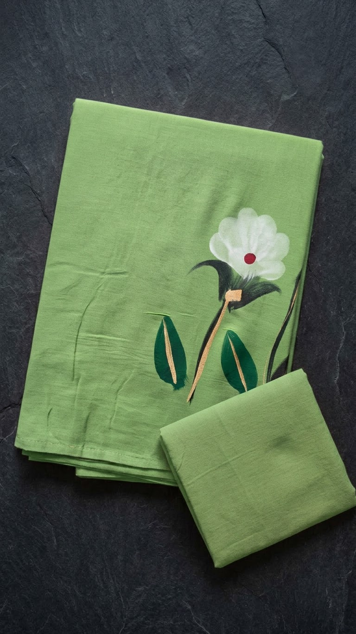 Light green cotton saree with a white flower, red centre, dark green leaves, and a plain light green blouse piece, folded on a charcoal background