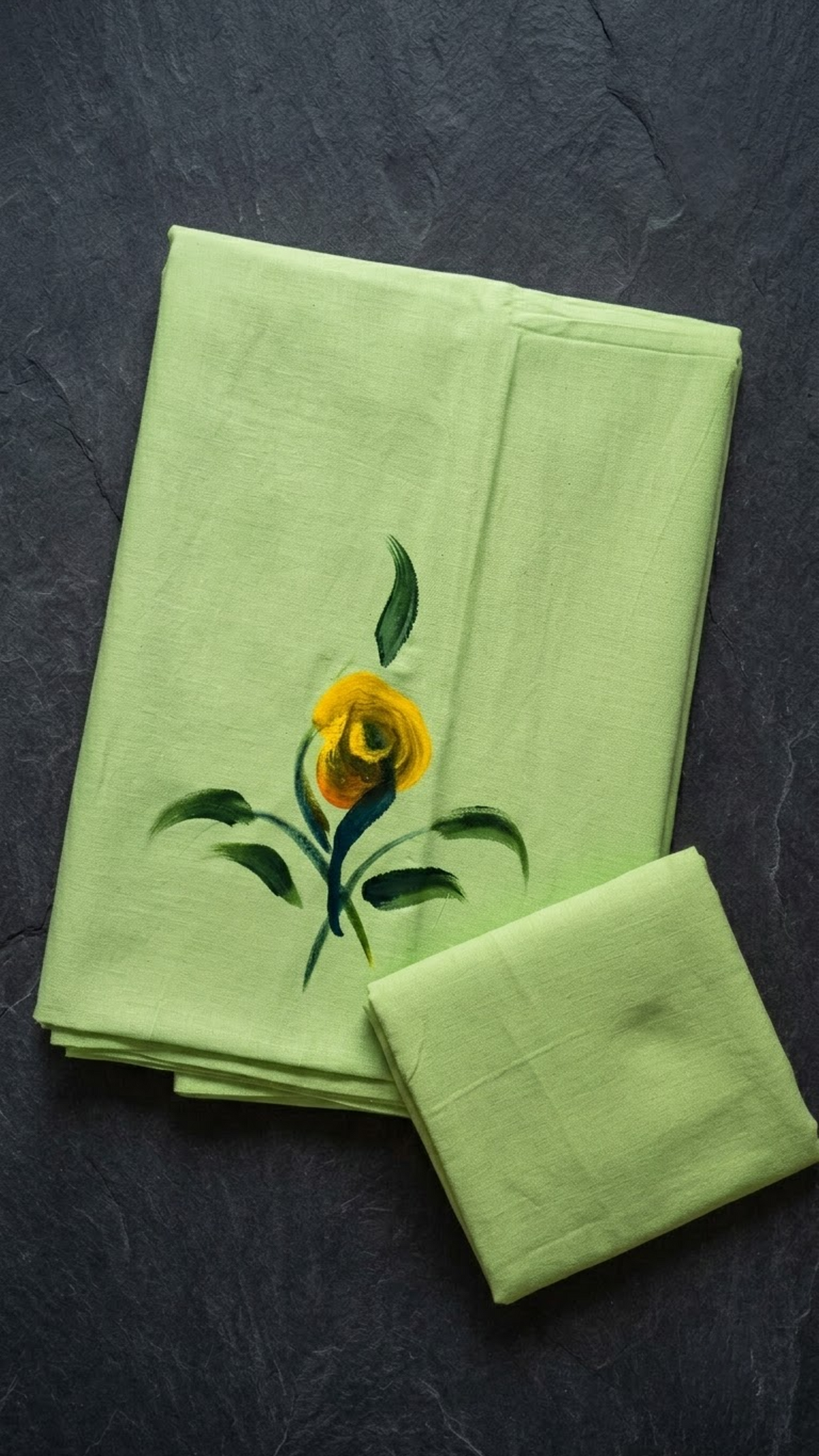 Light green cotton saree with a yellow flower, dark green leafy stems, and a plain light green blouse piece, folded on a charcoal background