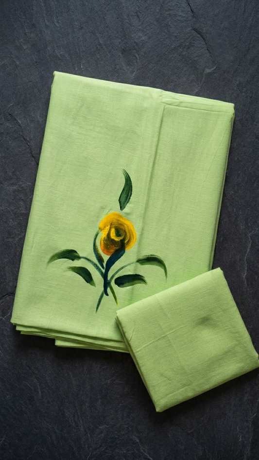 Light green cotton saree with a yellow flower, dark green leafy stems, and a plain light green blouse piece, folded on a charcoal background