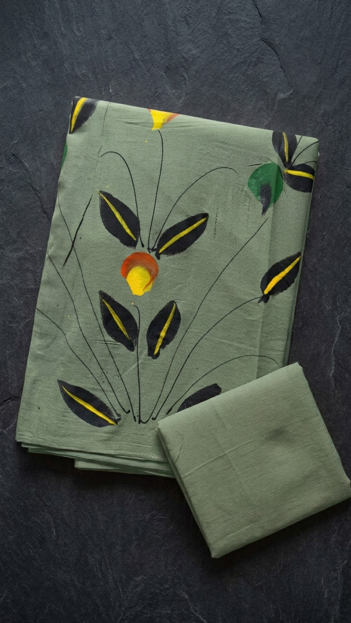 Sage green cotton saree with dark botanical leaves, yellow accents, and a plain sage green blouse piece, folded on a charcoal background