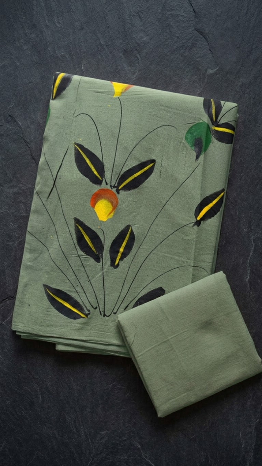 Sage green cotton saree with dark botanical leaves, yellow accents, and a plain sage green blouse piece, folded on a charcoal background
