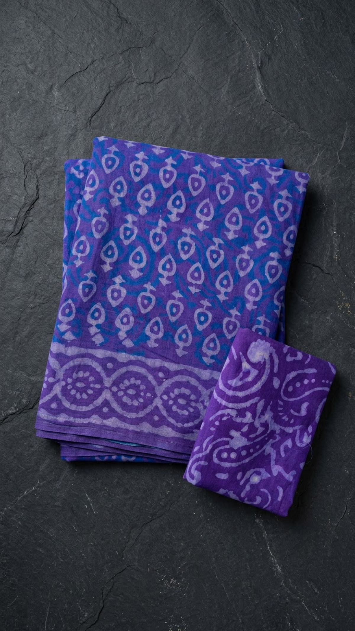 Purple cotton saree with light teardrop motifs, blue accents, a circular patterned border, and a purple paisley blouse piece, folded on a charcoal background