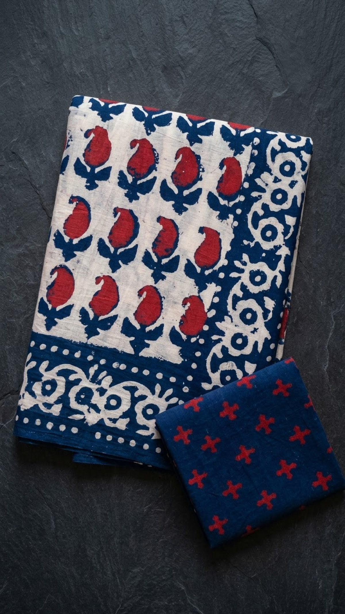 Ivory cotton saree with red buds, navy leaves, a navy-and-ivory floral border, and a navy red-cross blouse piece, folded on a charcoal background