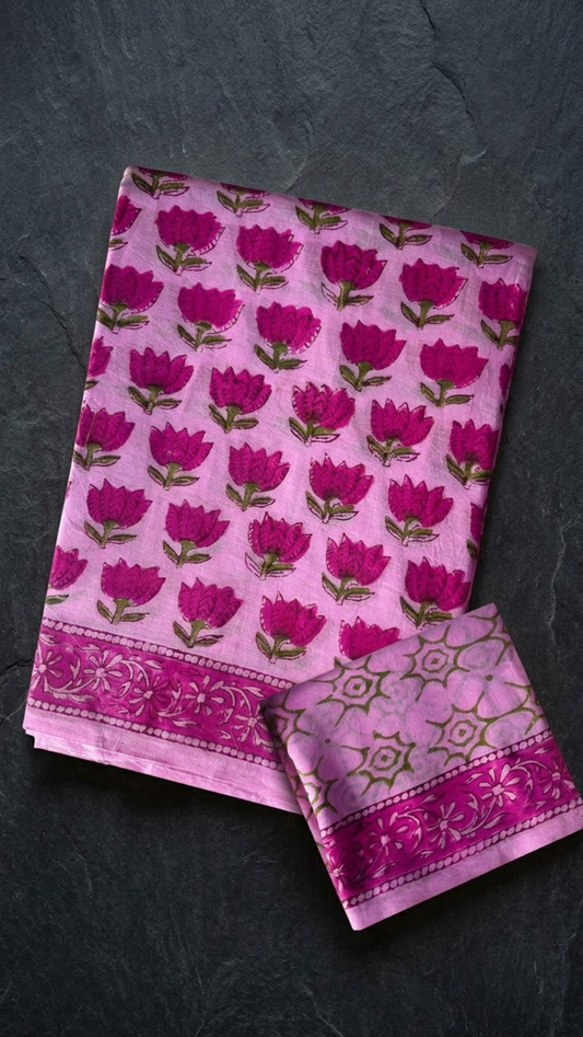 Pink cotton saree with magenta lotus motifs, a magenta floral border, and an olive geometric-print blouse piece, folded on a charcoal background