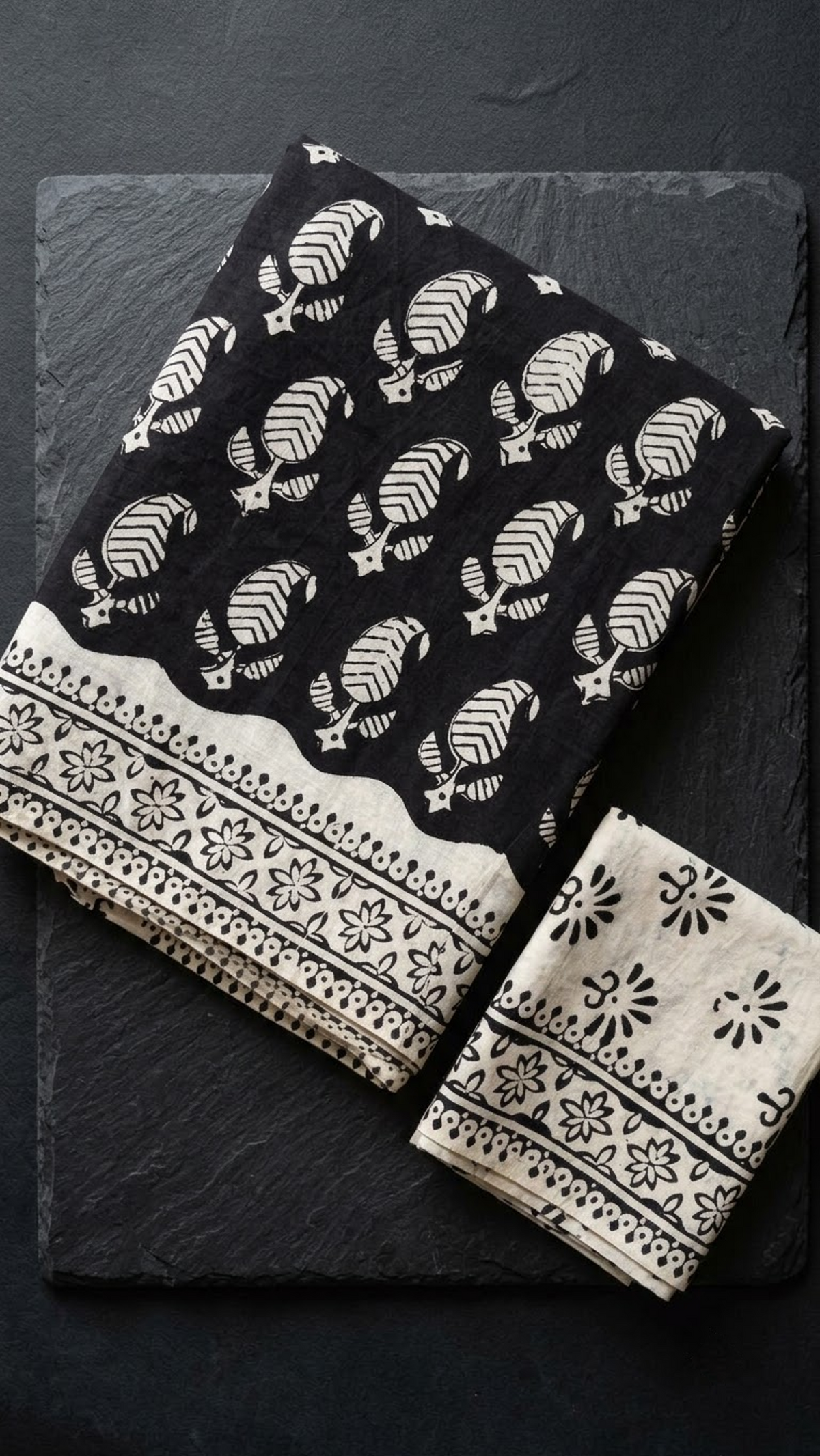 Black cotton saree with ivory leaf sprigs, an ivory floral border, and an ivory blouse piece with black floral motifs, folded on a charcoal background