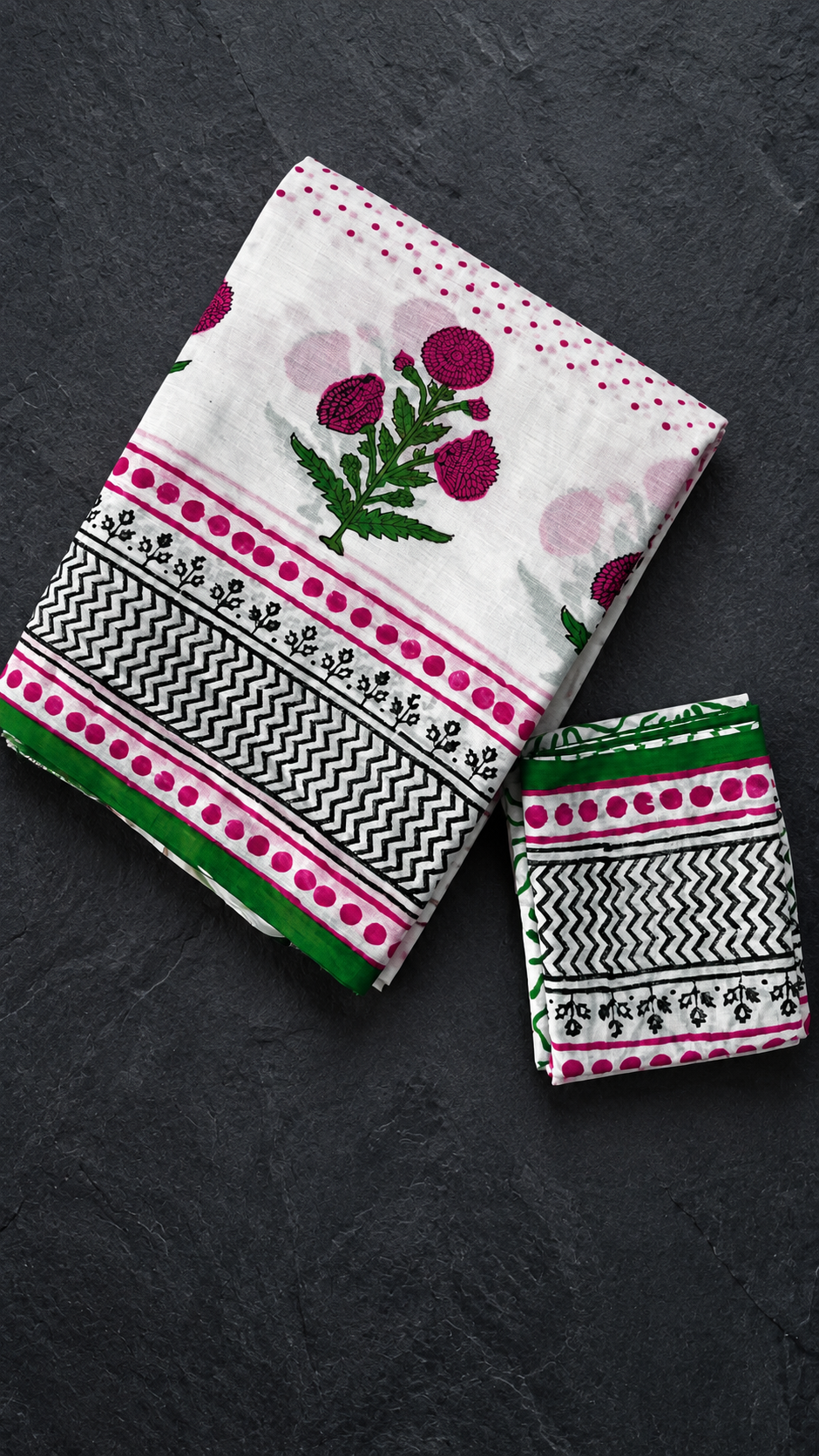 Ivory cotton saree with pink polka dots, a pink floral pallu, black zigzag border and green edging, folded on charcoal