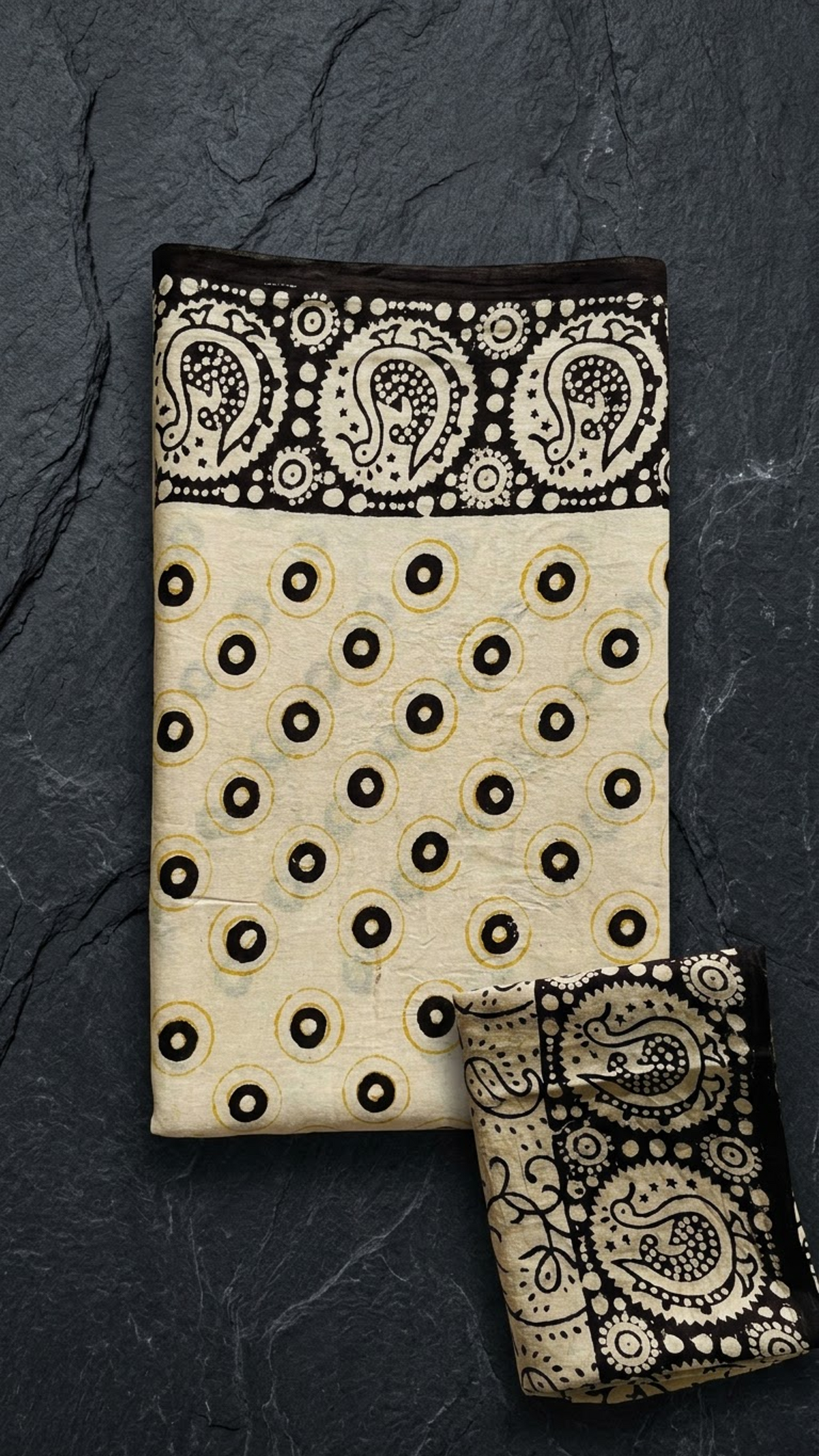 Ivory cotton saree with black circles and yellow rings, a black paisley border, and an ivory black vine-print blouse piece, folded on a charcoal background