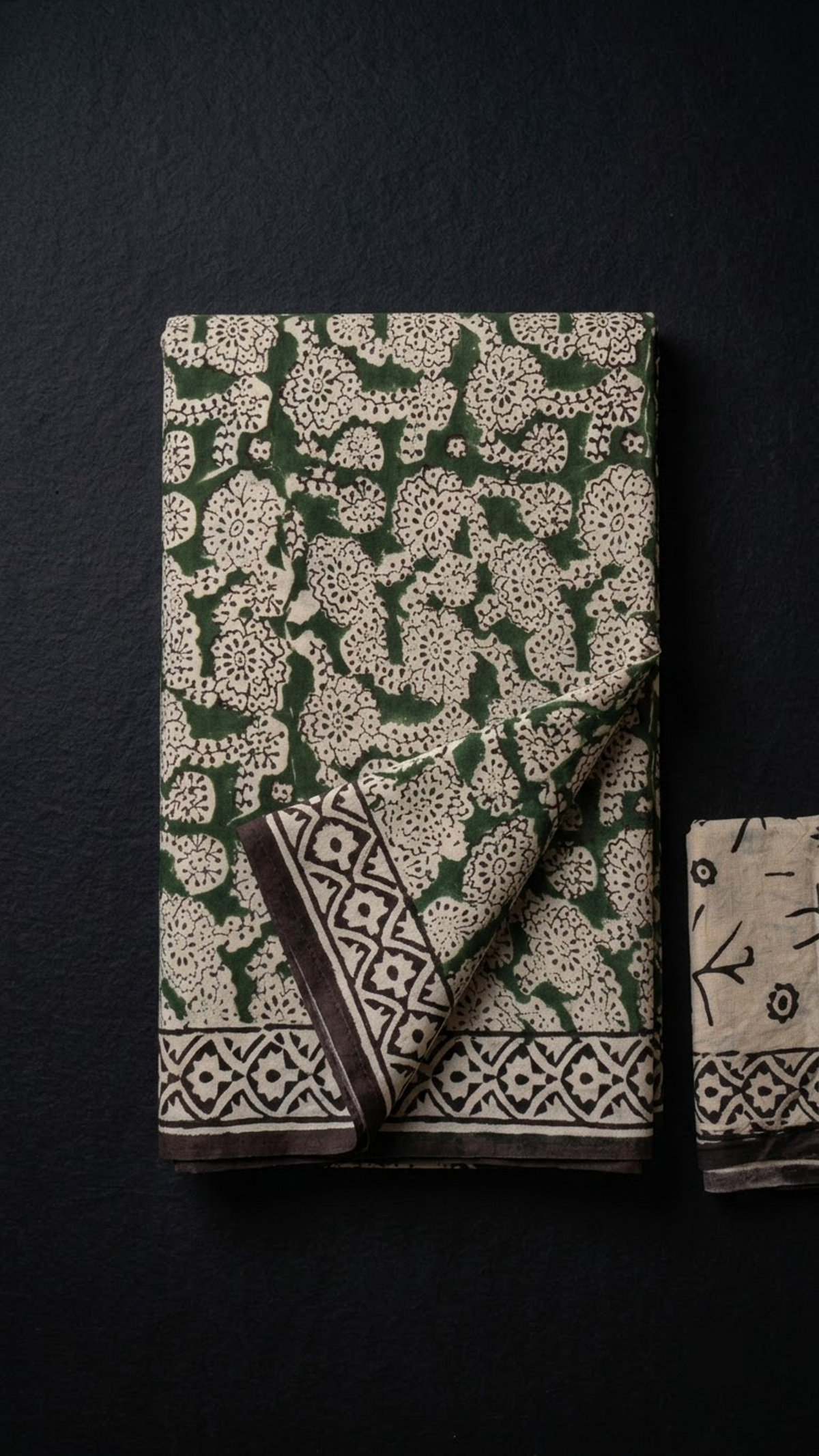 Folded green ornate floral cotton saree with blouse piece on a charcoal background