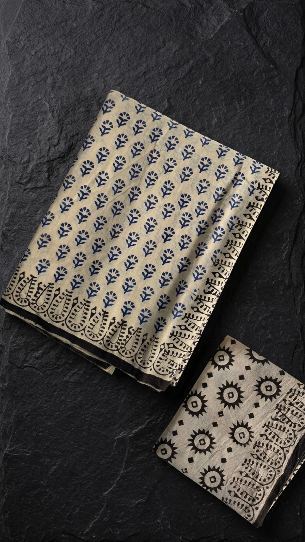 Ivory cotton saree with navy botanical sprigs, a black botanical loop border, and an ivory blouse piece with black starburst and diamond motifs, folded on a charcoal background