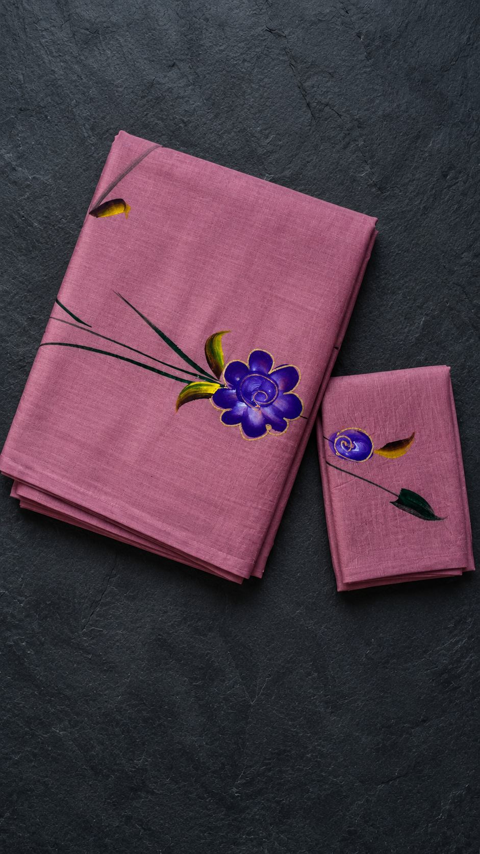 Mauve cotton saree with purple floral motifs, dark green stems, yellow-green leaves, and a matching mauve floral blouse piece, folded on a charcoal background