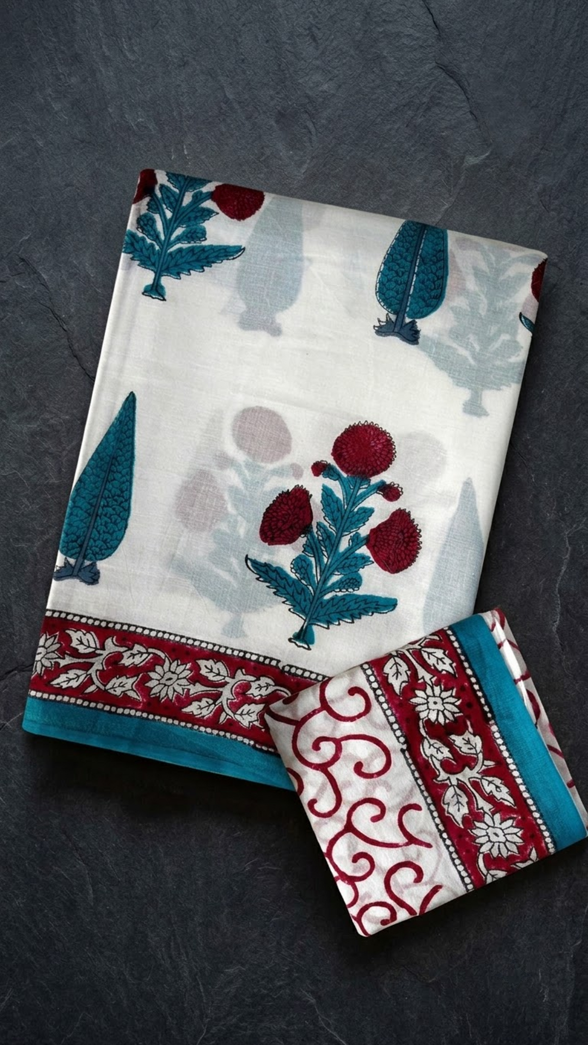 Ivory cotton saree with red flower clusters and teal foliage, a red floral border with teal edging, and an ivory red scroll-print blouse piece, folded on a charcoal background