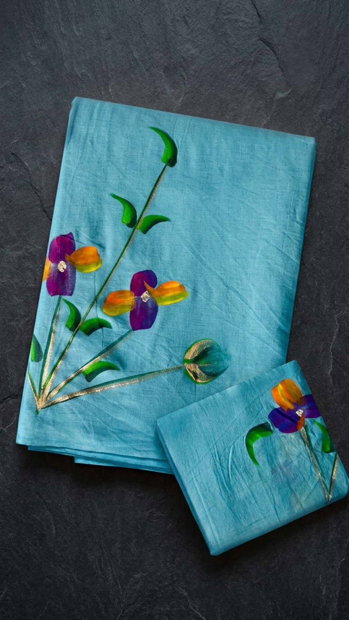 Turquoise cotton saree with purple and orange-yellow flowers, green stems, and a coordinating floral blouse piece, folded on a charcoal background