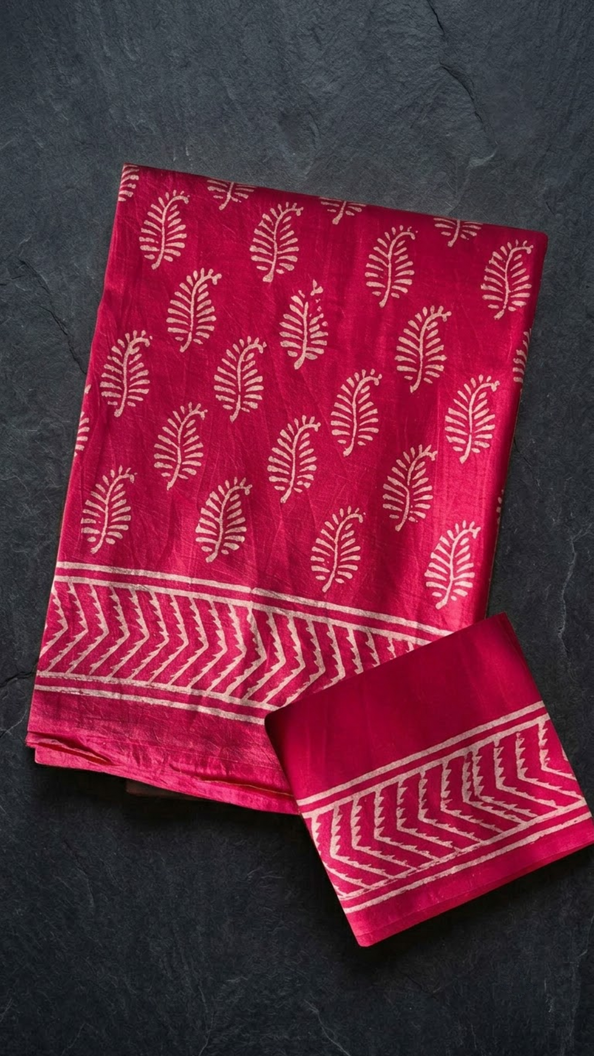 Deep pink cotton saree with ivory leafy sprigs, an ivory chevron border, and a matching bordered blouse piece, folded on a charcoal background