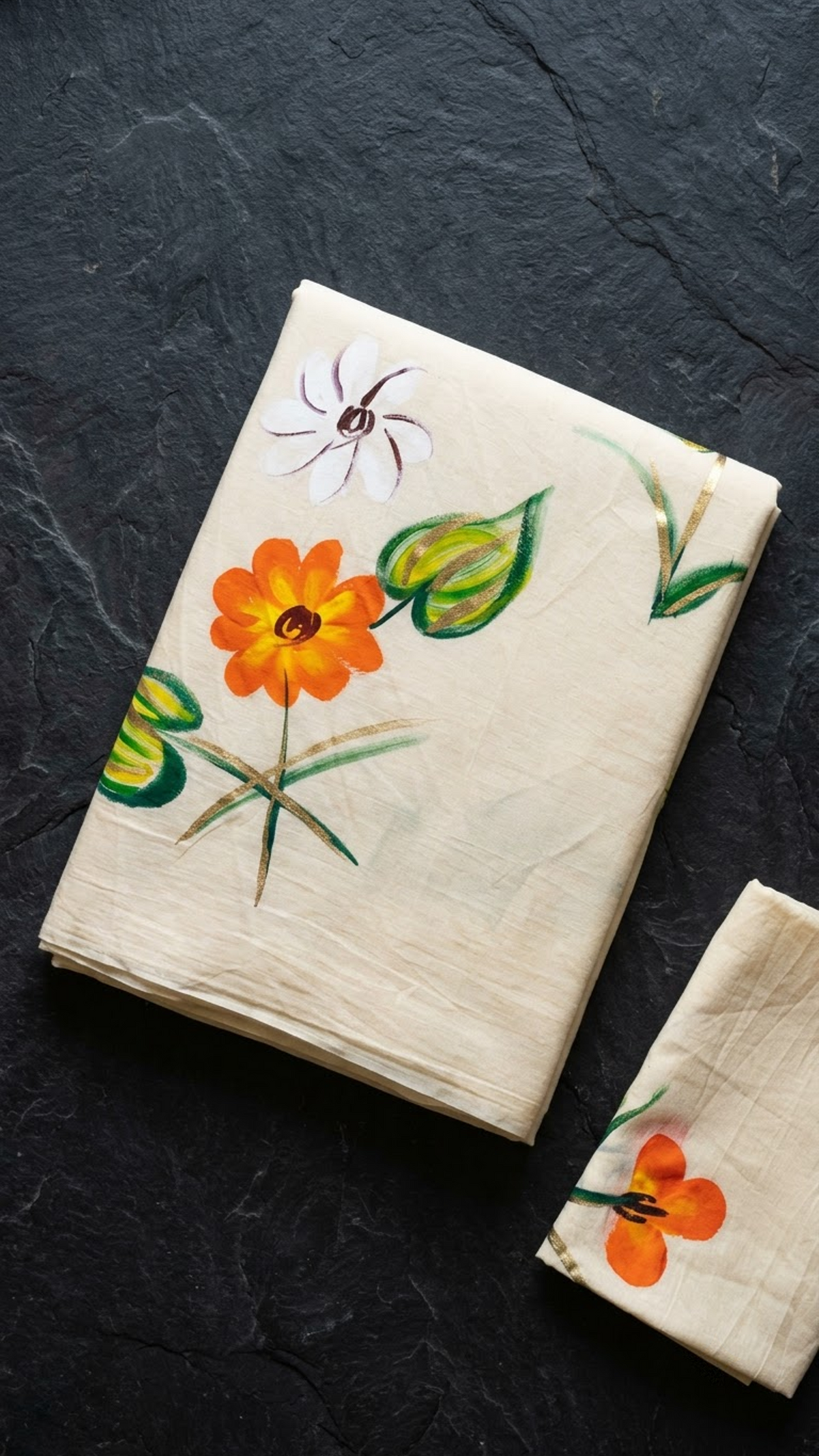 Ivory cotton saree with orange and white flowers, green leaves, and a coordinating orange floral blouse piece, folded on a charcoal background