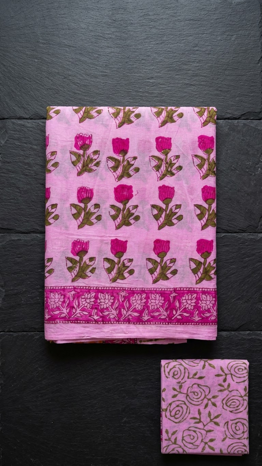 Pink cotton saree with magenta rosebuds, a magenta floral border, and a pink olive rose-print blouse piece, folded on a charcoal background