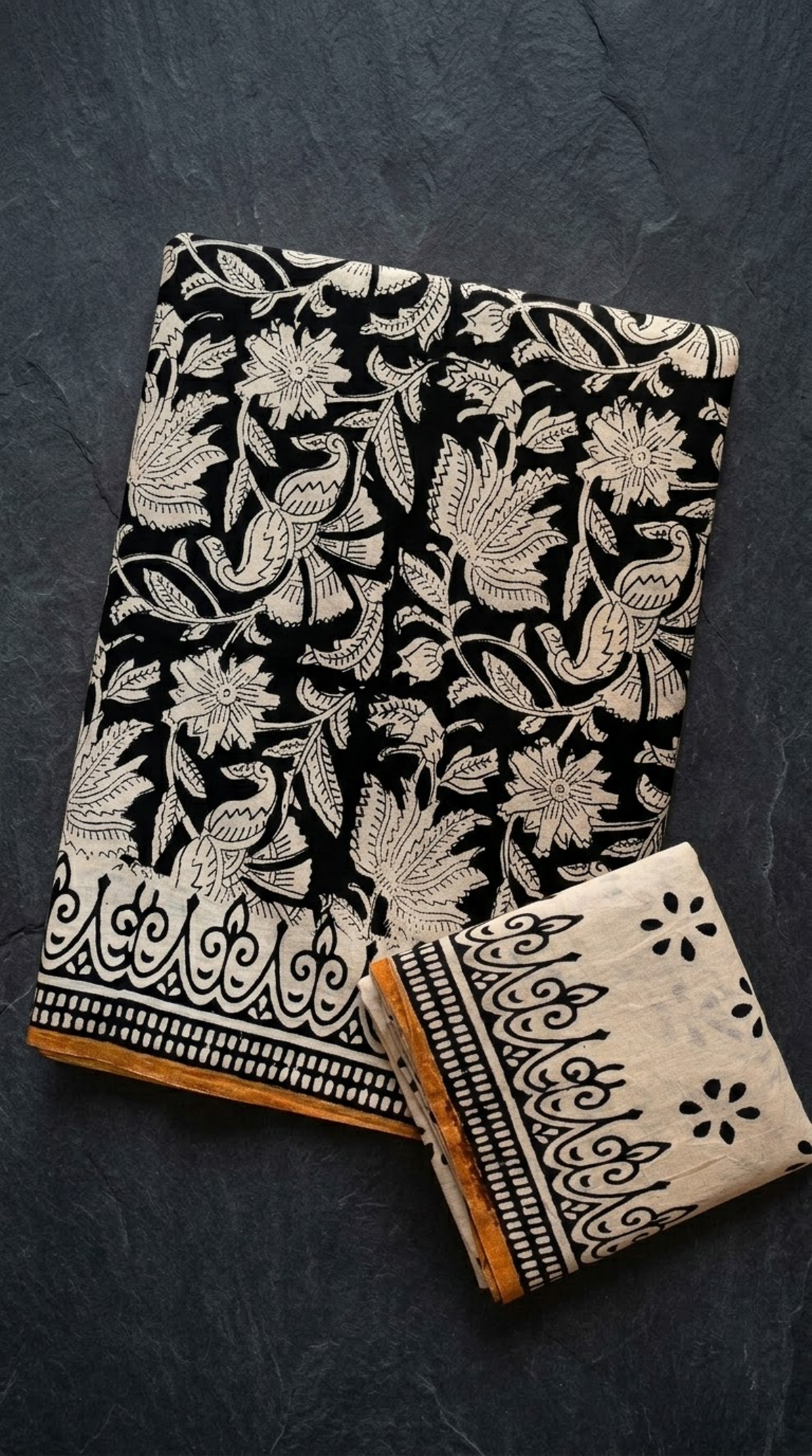 Black cotton saree with ivory bird and floral motifs, an ornamental border with an ochre edge, and an ivory blouse piece, folded on a charcoal background