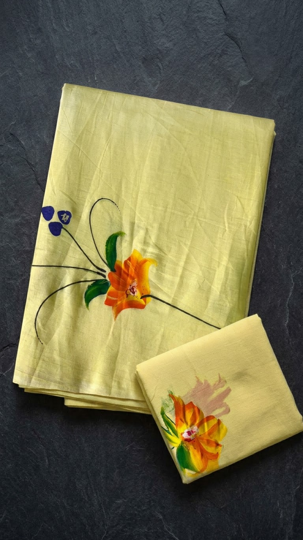 Lemon yellow cotton saree with an orange-yellow flower, green leaves, purple accents, and a coordinating floral blouse piece, folded on a charcoal background