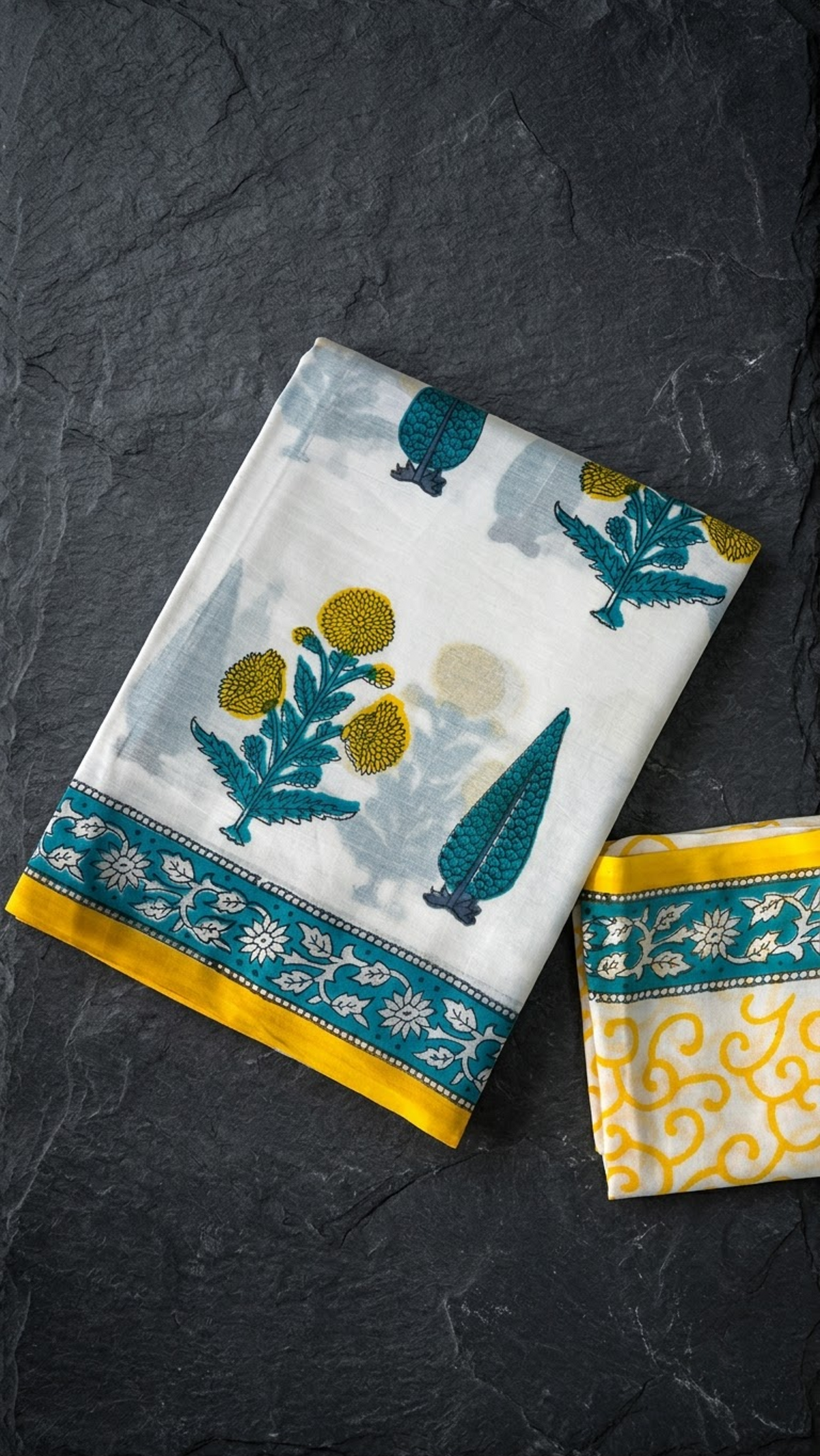 Ivory cotton saree with yellow flower clusters and teal foliage, a teal floral border with yellow edging, and an ivory yellow scroll-print blouse piece, folded on a charcoal background