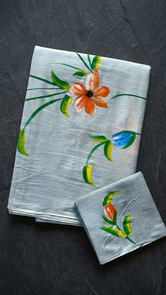 Pale blue cotton saree with an orange flower, blue bud, green stems, and a coordinating orange bud-print blouse piece, folded on a charcoal background