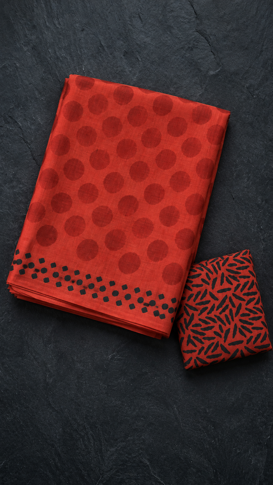 Rust red cotton saree with large tonal red polka dots, a border of small black geometric marks, and a black abstract print blouse piece, folded on a charcoal background